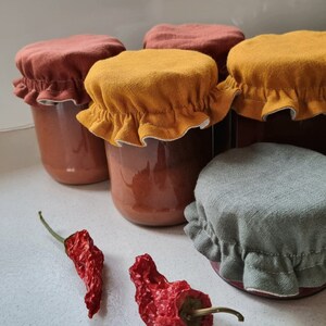 May include: Four glass jars with fabric lids in shades of brown, yellow, and gray. The jars contain a red sauce. Two dried red chili peppers are in the foreground.