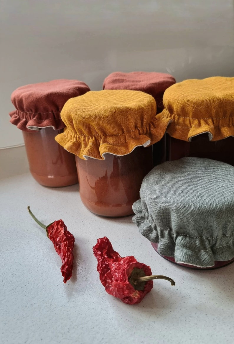 May include: Four glass jars with fabric lids in shades of brown, yellow, and gray. The jars contain a red sauce. Two dried red chili peppers are in the foreground.