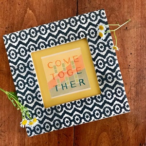 May include: A black and white patterned picture frame with a yellow mat and a colorful print inside that says "Come Together".