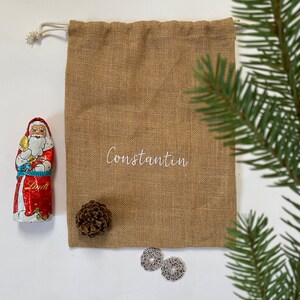 May include: A brown burlap gift bag with a white drawstring and the name "Constantin" embroidered on the front. The bag is filled with a chocolate Santa Claus, a pine cone, and two silver ornaments.