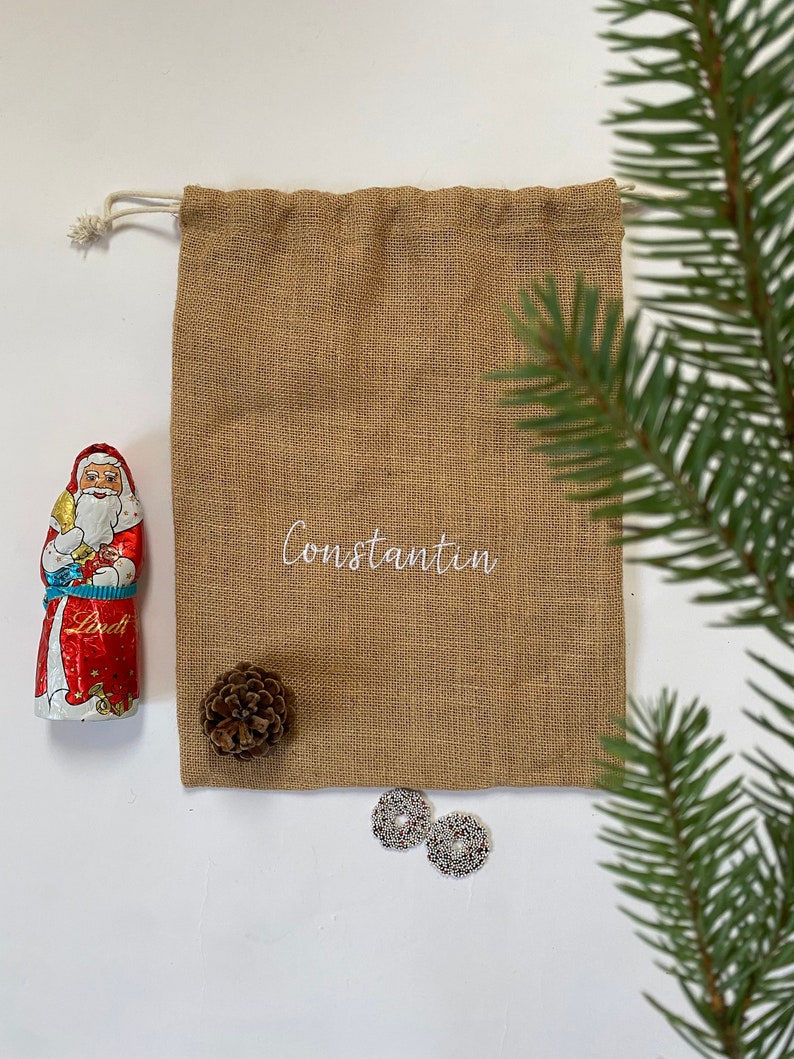 May include: A brown burlap gift bag with a white drawstring and the name "Constantin" embroidered on the front. The bag is filled with a chocolate Santa Claus, a pine cone, and two silver ornaments.
