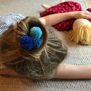 Könnte beinhalten: Eine junge Person mit langen blonden Haaren, die zu einem Knoten mit drei blauen und hellblauen Pompon-Haargummis gebunden sind. Die Person liegt auf einem hellgrauen Teppich.