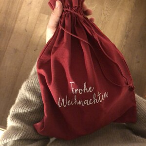 May include: A red fabric drawstring bag with white embroidery that says "Frohe Weihnachten" which is German for "Merry Christmas".