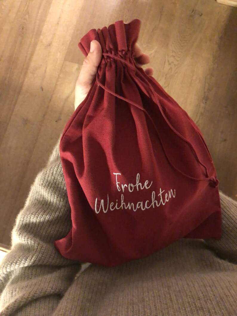 May include: A red fabric drawstring bag with white embroidery that says "Frohe Weihnachten" which is German for "Merry Christmas".