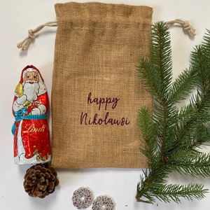 May include: A burlap gift bag with a drawstring closure, decorated with the text "happy Nikolaus" in maroon lettering. The bag is filled with a chocolate Santa Claus figurine, a pine cone, and two white and silver beaded ornaments.
