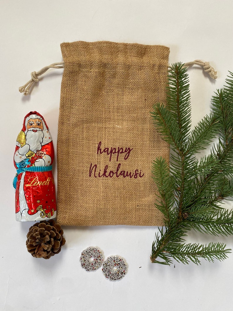 May include: A burlap gift bag with a drawstring closure, decorated with the text "happy Nikolaus" in maroon lettering. The bag is filled with a chocolate Santa Claus figurine, a pine cone, and two white and silver beaded ornaments.