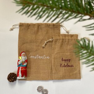 May include: Two brown burlap gift bags with white drawstring closures. The larger bag has the name "Constantin" embroidered on it in white thread. The smaller bag has the text "happy Nikolaus" embroidered on it in red thread.