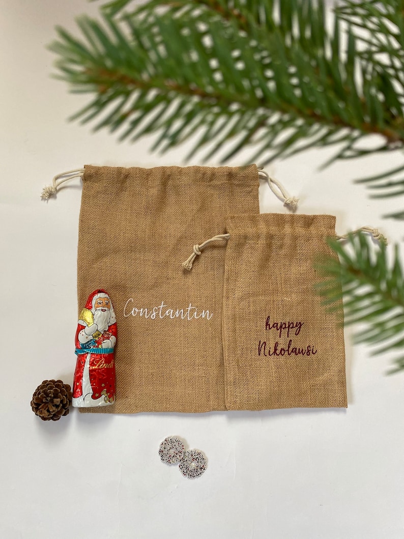 May include: Two brown burlap gift bags with white drawstring closures. The larger bag has the name "Constantin" embroidered on it in white thread. The smaller bag has the text "happy Nikolaus" embroidered on it in red thread.