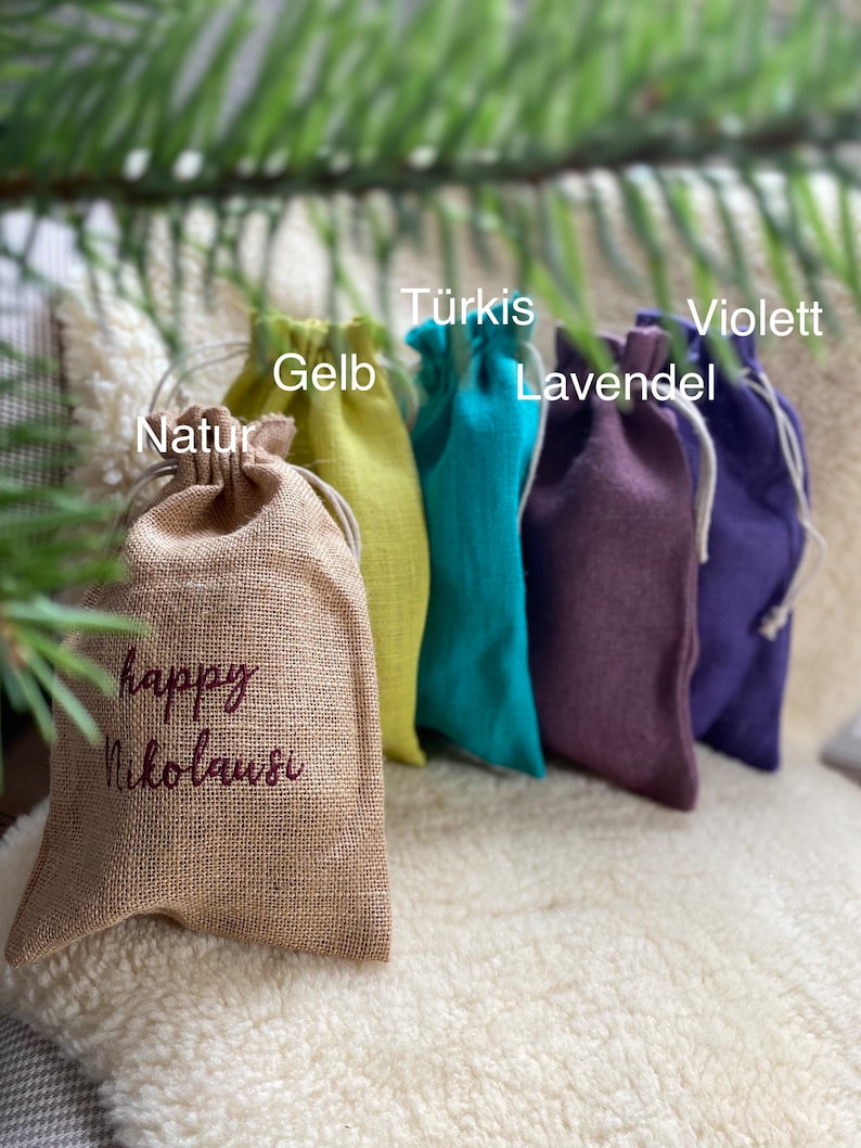 May include: Five small fabric gift bags in different colors. The bags are lined up from left to right: natural, yellow, turquoise, purple, and violet. The bag on the left has the text "happy Nikolaus" embroidered on it.