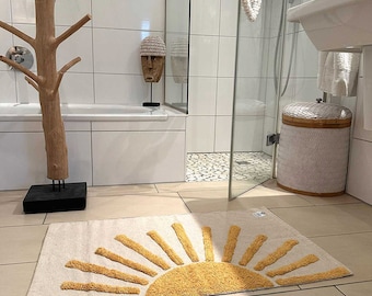 May include: A beige bath mat with a yellow sun design. The mat is in a bathroom with white tile walls and a white bathtub.