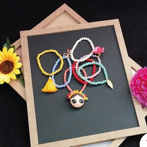 Puede incluir: Una colecci&oacute;n de collares y pulseras de cuentas coloridas dispuestas sobre una superficie negra. Los collares presentan cuentas de varios colores, incluyendo amarillo, rojo, azul y blanco, con un colgante central de una cara de dibujos animados. Tambi&eacute;n se ve una borla amarilla y un dije de hoja dorada.