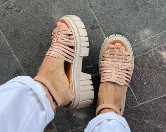 May include: A pair of beige woven platform sandals with ankle straps. The sandals are worn by a person wearing white pants.