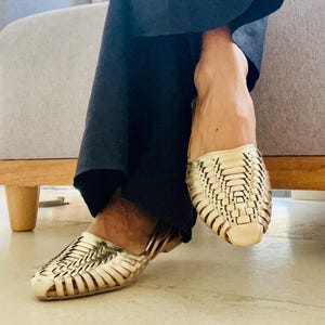 May include: Gold woven leather sandals. The sandals have a flat sole and a woven design across the top. The sandals are worn with dark blue pants.