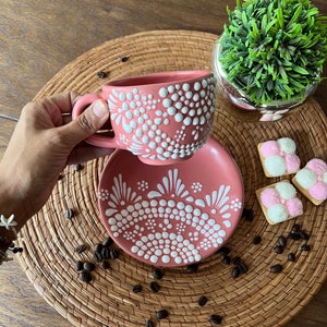 May include: A pink ceramic teacup and saucer with a white dot and floral design. The cup and saucer are on a woven placemat with coffee beans.