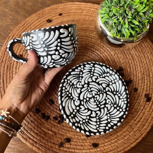 May include: A black and white ceramic coffee cup and saucer set with a swirling floral pattern. The cup and saucer are on a woven placemat with coffee beans scattered around them.