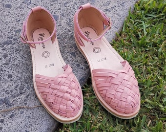 May include: A pair of pink leather sandals with woven straps and an ankle strap with a buckle. The sandals have the brand name "Macapa" printed on the straps.