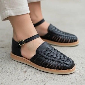 May include: Black leather sandals with woven detailing on the vamp and a small ankle strap with a gold-colored buckle. The sandals have a light brown sole and are worn by a person wearing light-colored pants.