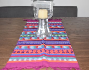 Op de afbeelding: Een kleurrijke tafelrunner met een geometrisch patroon in tinten roze, oranje, blauw, groen en paars. De runner is gemaakt van geweven stof en heeft franjes aan de uiteinden. Een zilveren kaarsenhouder met een transparante glazen bovenkant staat op de runner.