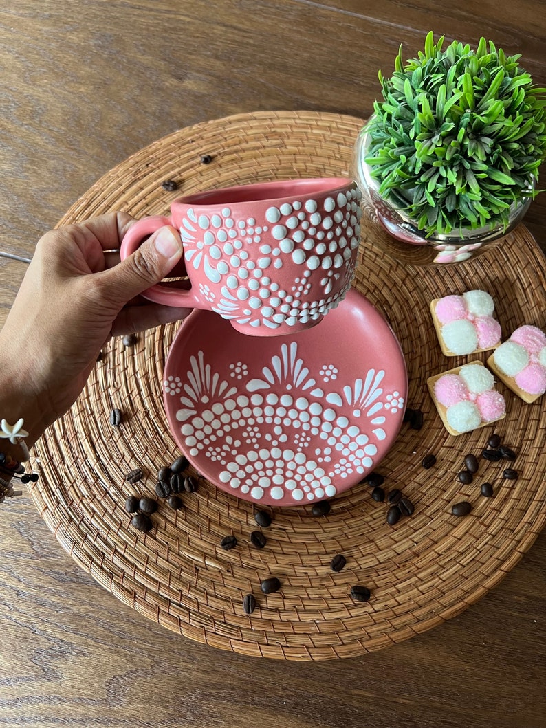 May include: A pink ceramic teacup and saucer with a white dot and floral design. The cup and saucer are on a woven placemat with coffee beans.