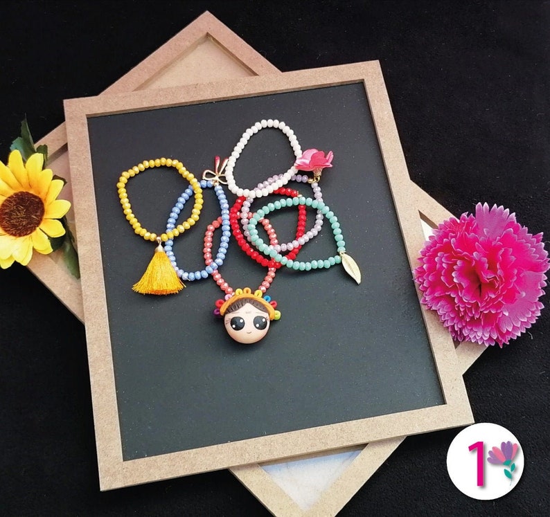 Puede incluir: Una colecci&oacute;n de collares y pulseras de cuentas coloridas dispuestas sobre una superficie negra. Los collares presentan cuentas de varios colores, incluyendo amarillo, rojo, azul y blanco, con un colgante central de una cara de dibujos animados. Tambi&eacute;n se ve una borla amarilla y un dije de hoja dorada.