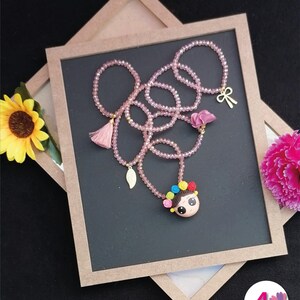 Puede incluir: Un collar de cuentas rosas con un colgante con la cara de una ni&ntilde;a de dibujos animados, adornado con detalles florales coloridos. El collar incluye un dije de lazo dorado, una borla y un dije de hoja. El collar se exhibe sobre una superficie gris oscuro.