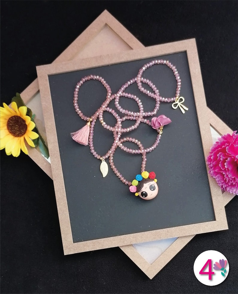 Puede incluir: Un collar de cuentas rosas con un colgante con la cara de una ni&ntilde;a de dibujos animados, adornado con detalles florales coloridos. El collar incluye un dije de lazo dorado, una borla y un dije de hoja. El collar se exhibe sobre una superficie gris oscuro.