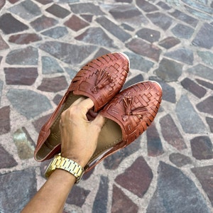 May include: A pair of brown leather sandals with woven straps and tassels. The sandals are held in a person's hand.