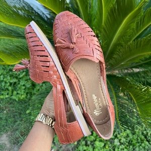 May include: A pair of brown leather sandals with a woven design and tassel accents. The sandals have a white sole and the brand name "Macarena" is printed on the insole.