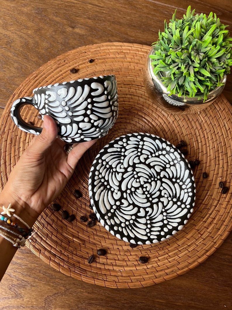 May include: A black and white ceramic coffee cup and saucer set with a swirling floral pattern. The cup and saucer are on a woven placemat with coffee beans scattered around them.