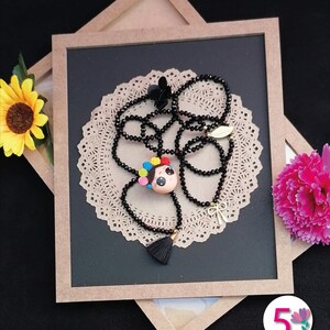Puede incluir: Un collar hecho a mano con una cadena de cuentas negras y un colgante con forma de cara de mu&ntilde;eca con pelo colorido. El collar incluye una borla negra y un dije de lazo dorado. Se exhibe sobre un mantelito de papel decorativo.