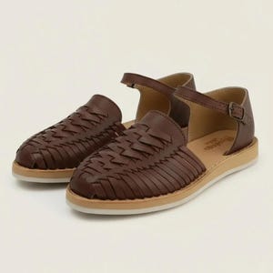 May include: A pair of brown leather sandals with woven detailing. The sandals have a closed-toe design with a strap and buckle closure. The soles are a light tan colour.