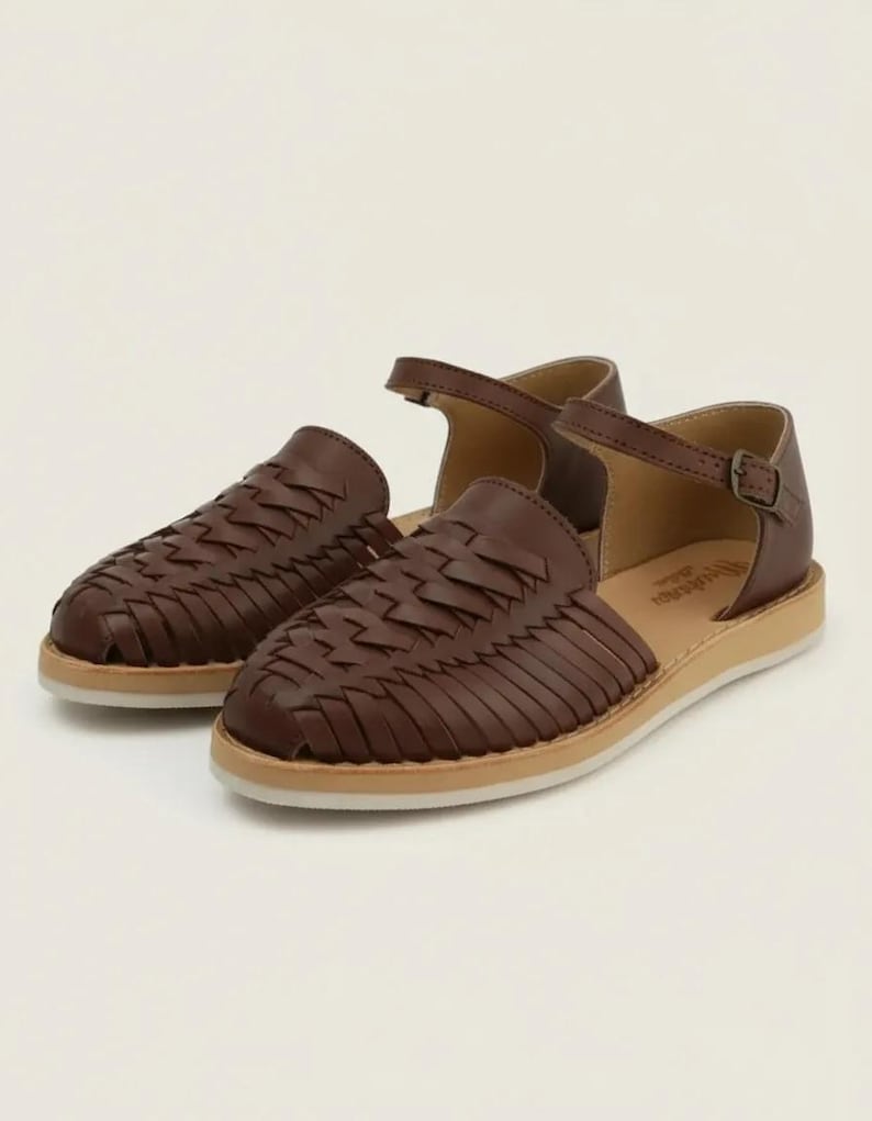 May include: A pair of brown leather sandals with woven detailing. The sandals have a closed-toe design with a strap and buckle closure. The soles are a light tan colour.