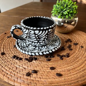 May include: A black and white ceramic coffee cup and saucer with a floral design. The cup and saucer are sitting on a woven placemat with coffee beans scattered around them.