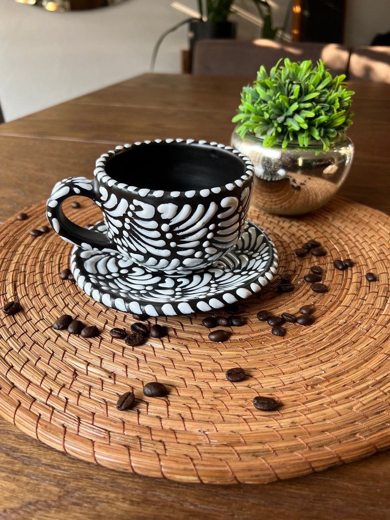 May include: A black and white ceramic coffee cup and saucer with a floral design. The cup and saucer are sitting on a woven placemat with coffee beans scattered around them.
