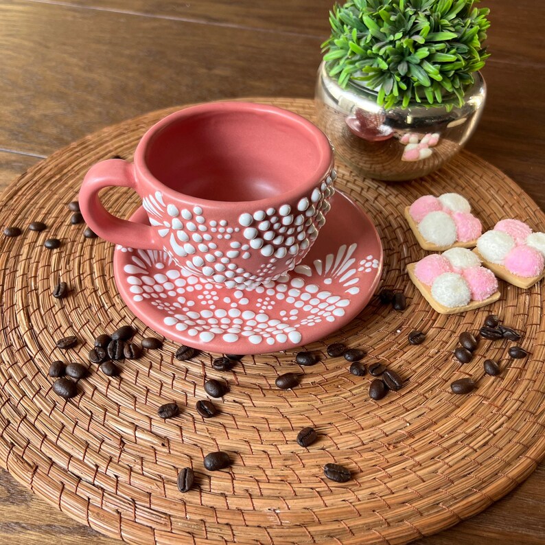 May include: A pink ceramic coffee cup and saucer with a white polka dot design. The cup and saucer are sitting on a woven placemat with coffee beans scattered around them.