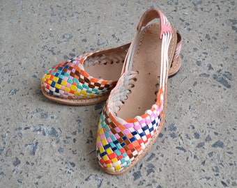 May include: A pair of huarache sandals featuring a multi-colored woven leather upper with a vibrant pattern of pink, orange, yellow, blue, and brown squares. The sandals have a tan leather footbed, a slingback strap, and a flat sole construction.