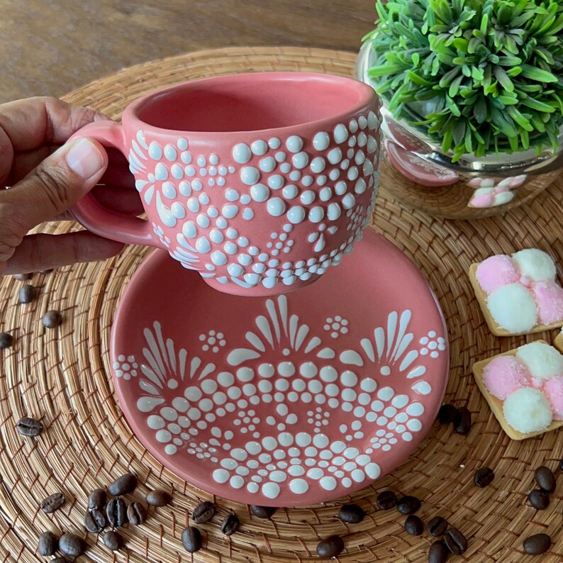 May include: A pink teacup and saucer set with a white polka dot and floral design. The set is on a woven placemat with coffee beans scattered around it.