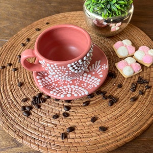 May include: A pink ceramic coffee cup and saucer with a white polka dot design. The cup and saucer are on a woven placemat with coffee beans scattered around them.