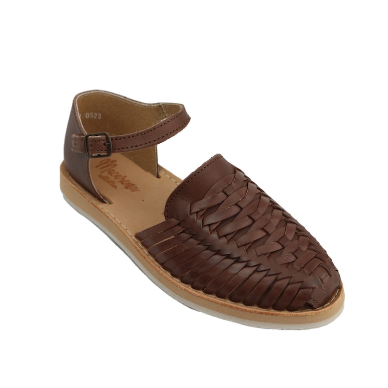 May include: Brown leather huarache sandal with a woven vamp and ankle strap. The sandal has a buckle fastening and a light-coloured sole. The brand name is visible on the side. The sandal is designed for casual wear.