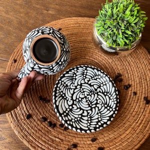 May include: Black and white ceramic coffee cup and saucer set with a swirling pattern. The cup is filled with coffee and the saucer is on a woven mat.