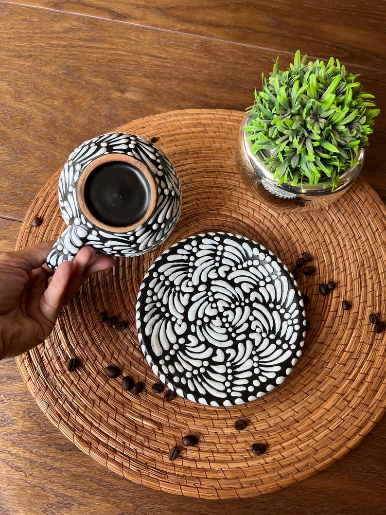 May include: Black and white ceramic coffee cup and saucer set with a swirling pattern. The cup is filled with coffee and the saucer is on a woven mat.