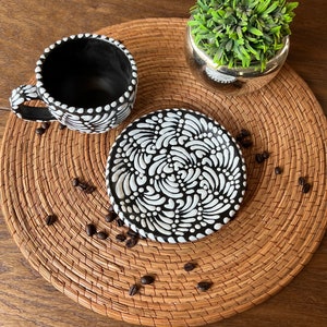 May include: A black and white ceramic coffee cup and saucer set with a decorative pattern. The cup and saucer are on a woven placemat with coffee beans scattered around them.