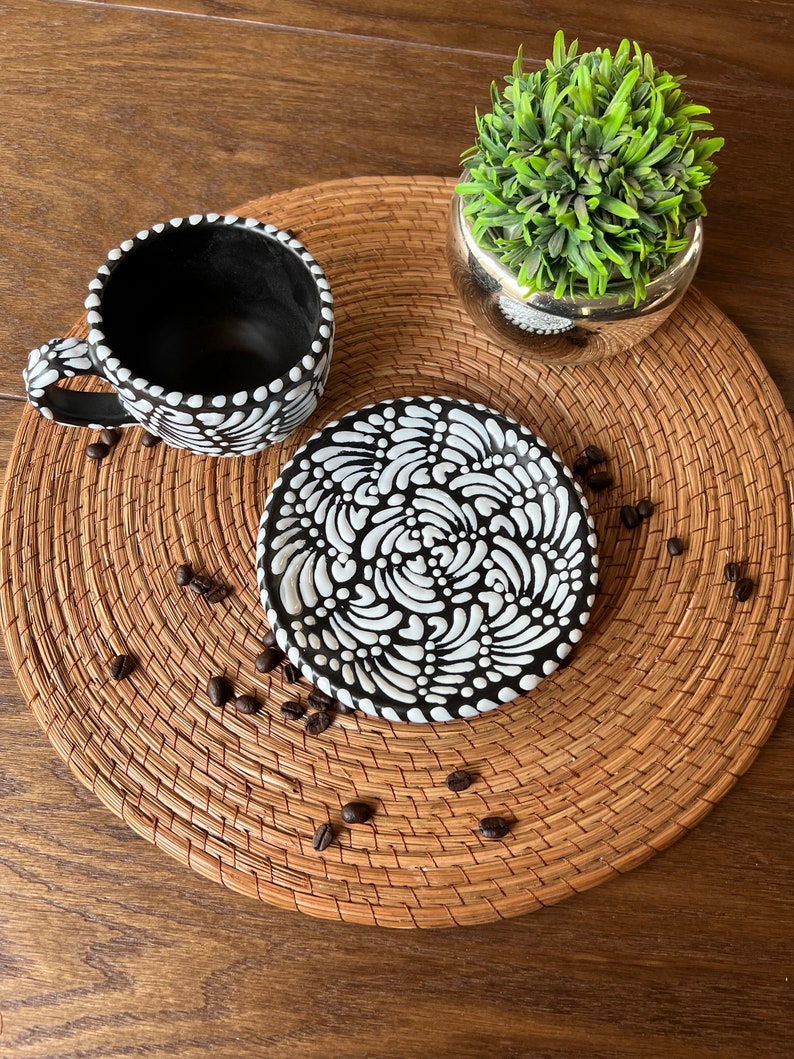 May include: A black and white ceramic coffee cup and saucer set with a decorative pattern. The cup and saucer are on a woven placemat with coffee beans scattered around them.