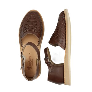 May include: Brown leather sandals with woven detailing and a buckle fastening. The sandals feature a light-coloured sole and an ankle strap. The brand name "Macarena collection" is printed on the insole.