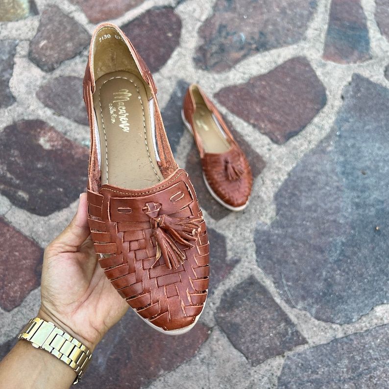 May include: A pair of brown leather sandals with a woven design and tassel accents. The sandals are on a stone surface.