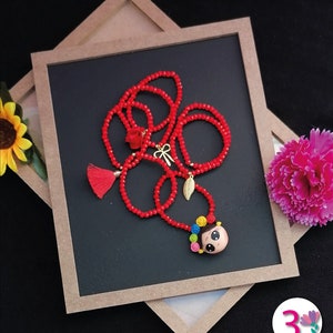 Puede incluir: Un collar de cuentas rojas con un colgante que presenta a una ni&ntilde;a de dibujos animados con flores de colores en el pelo. El collar incluye una borla y abalorios dorados. El collar se exhibe sobre una superficie negra.