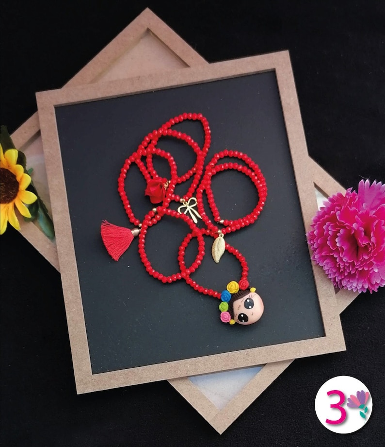 Puede incluir: Un collar de cuentas rojas con un colgante que presenta a una ni&ntilde;a de dibujos animados con flores de colores en el pelo. El collar incluye una borla y abalorios dorados. El collar se exhibe sobre una superficie negra.