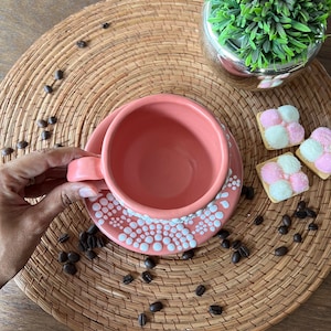 May include: A pink ceramic coffee cup with white polka dots on the rim and saucer. The cup and saucer are on a woven placemat with coffee beans scattered around them.