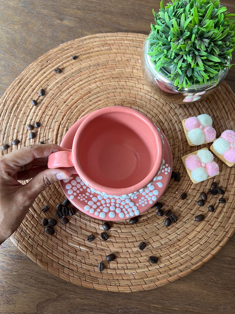 May include: A pink ceramic coffee cup with white polka dots on the rim and saucer. The cup and saucer are on a woven placemat with coffee beans scattered around them.