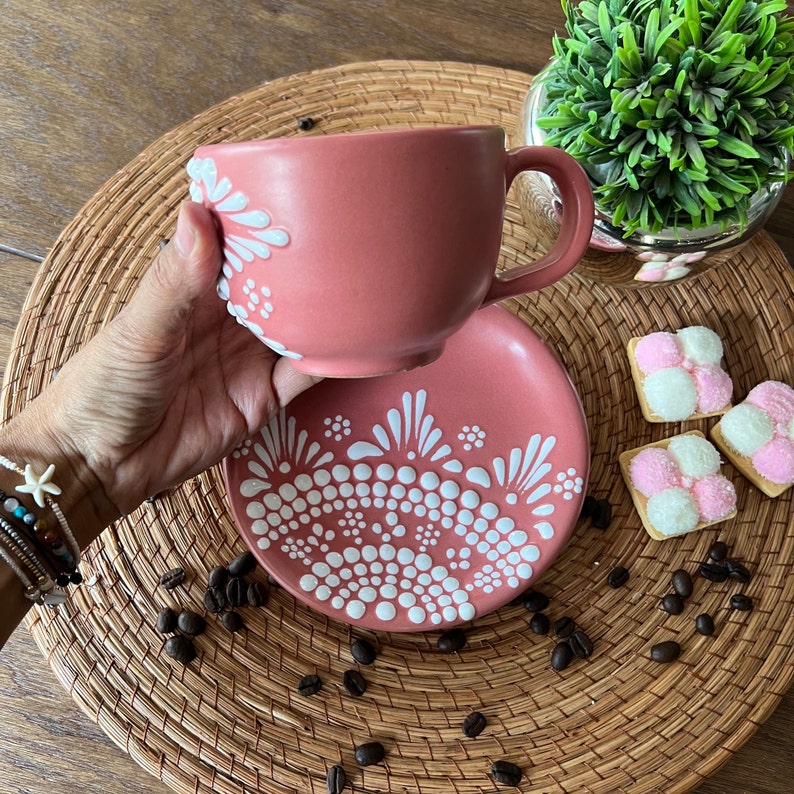 May include: A pink ceramic teacup and saucer with a white dot and swirl design. The cup and saucer are on a woven placemat with coffee beans scattered around them.